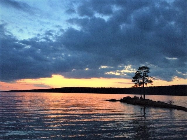 “A small island with one tall tree is surrounded by lake water. The sun has set behind a mountain and clouds cover half of the sky.” 